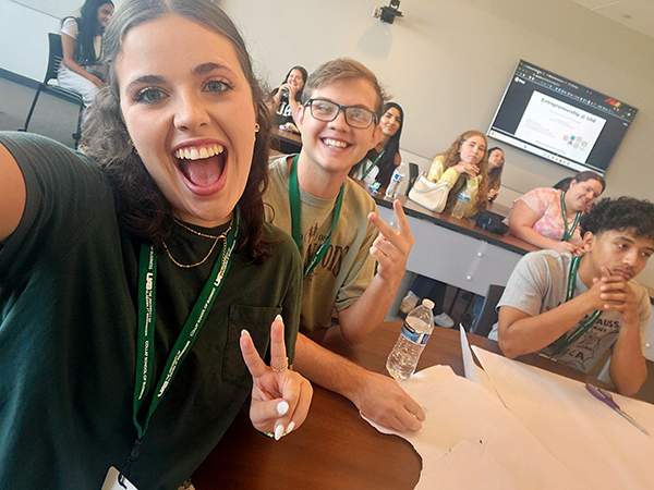 two students in green smiling at the camera and showing peace signs with their fingers