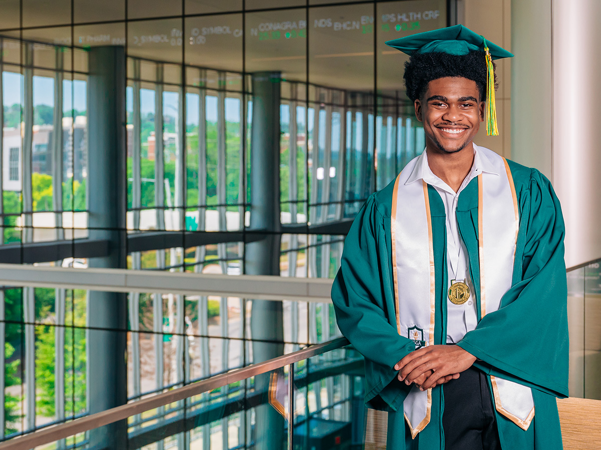 A smiling student with short black hair wearing a green graduation robe with a white stole and a green mortarboard with a gold tassel