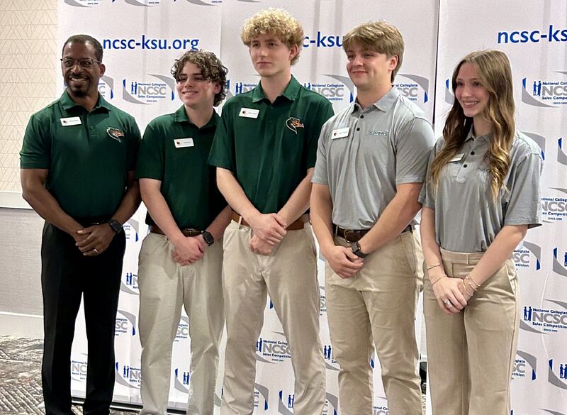 a professor and four student stand in front of a backdrop, hands clasped in front of them, and smile at a camera out of shot