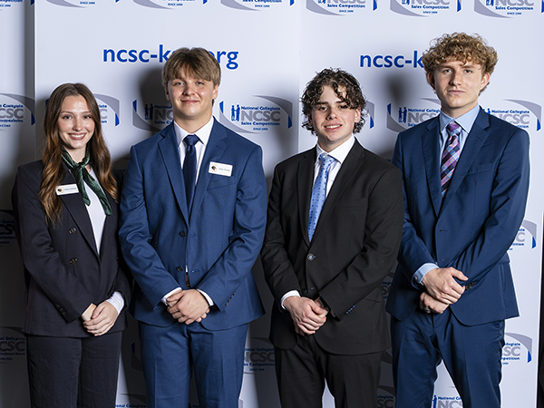 four students wearing dark suits and ties standing side by side, smiling with hands clasped in front of them