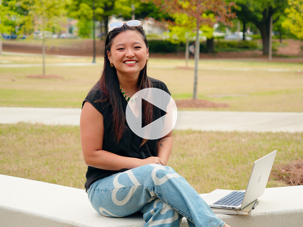Smiling student with long dark hair, glasses on top of her head, wearing light colored jeans with hearts on the, sitting outdoors next to an open laptop