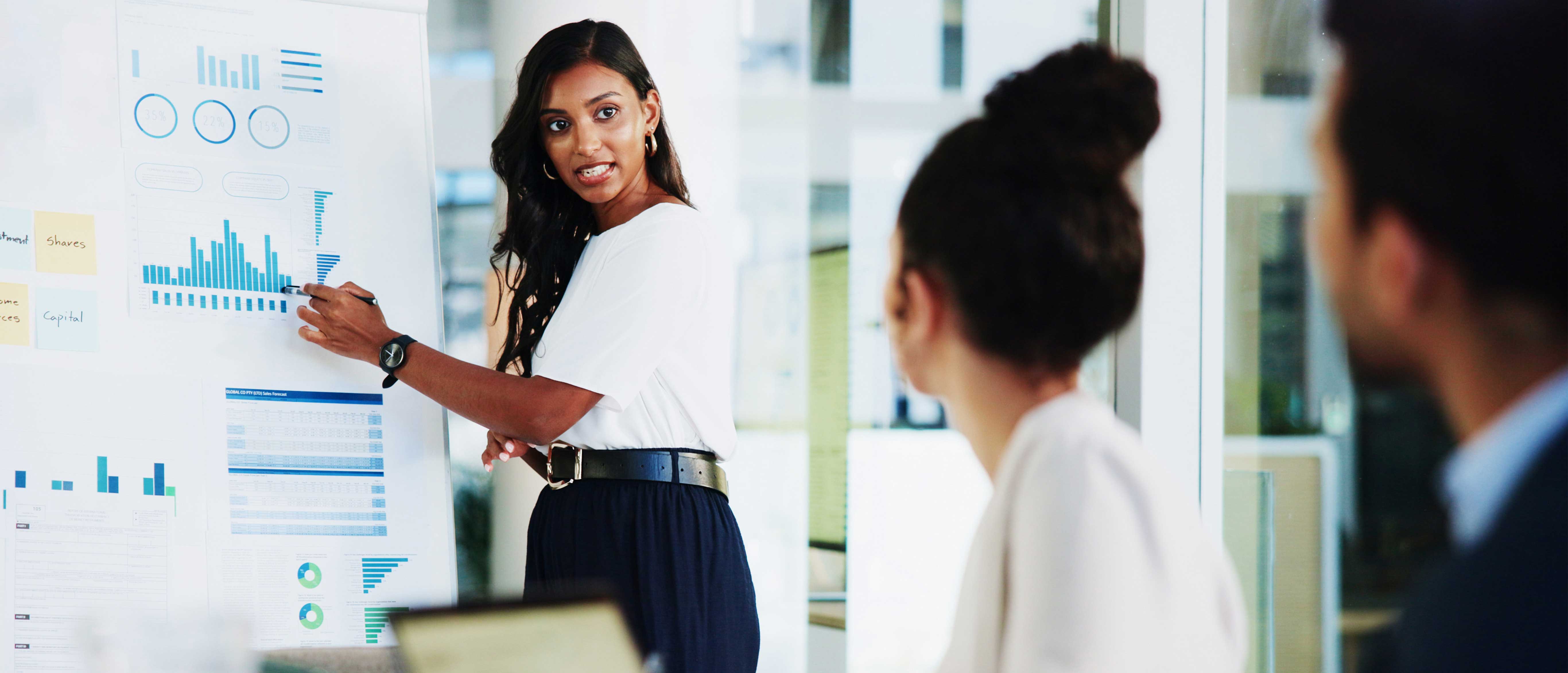 Woman presenting data charts on a whiteboard to two colleagues in an office meeting.
