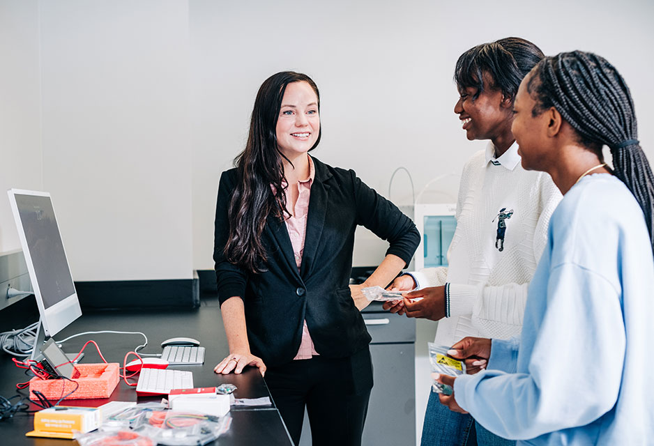 A teacher talks to two female students in a classrom next two a computer and many components. 