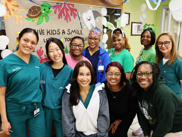 Group photo of faculty, staff and students standing in front of the Give Kids a Smile poster in the UAB School of Dentistry pediatric clinic.