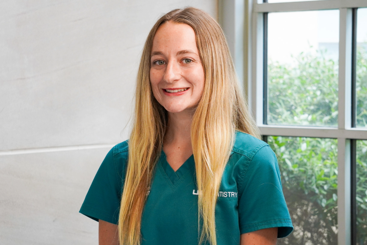 Blonde woman earing green scrubs smiling for a photo with a concrete wall and window in the background