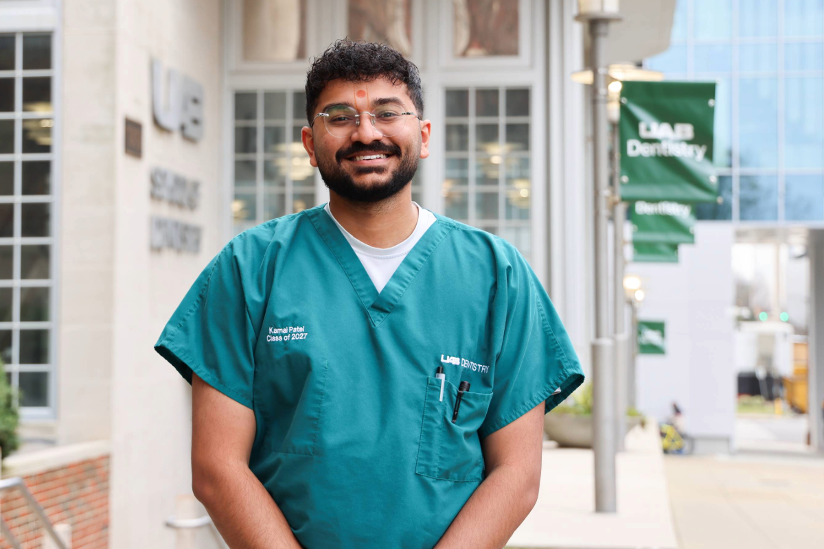 Male dental student (Kamal Patel) smiling wearing green scrubs in front of the UAB School of Dentistry building