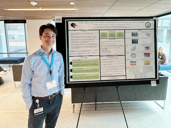 Ethan Brenna (white male in blue button up and slacks) smiling while presenting their research poster on pediatric craniofacial trauma at SEMSS 2025.