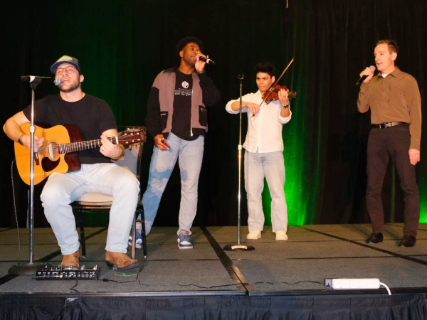 Three students and faculty member on stage at the 2026 Variety Show (Left to right: Seth Killam is sitting while playing guitar and singing, Josh Holsey is standing while singing, Edwin Rojas is standing while playing violin, and Dr. Goodspeed is standing while singing)