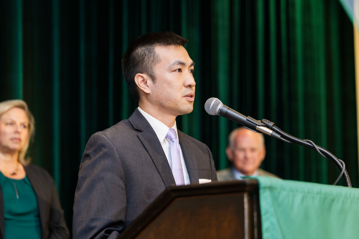 Dr. Liu, a man in a suit speaking at a podium at award ceremony with others in the background