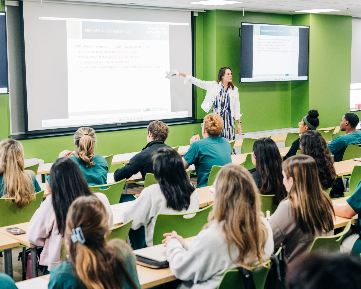 Woman faculty member pointing towards board in classroom full of students in green scrubs