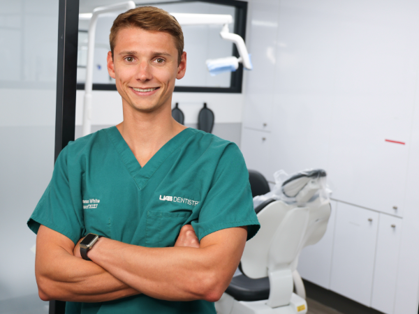 Young dental student in green scrubs smiling with arms crossed in a bright, modern dental operatory beside a patient chair and overhead light.