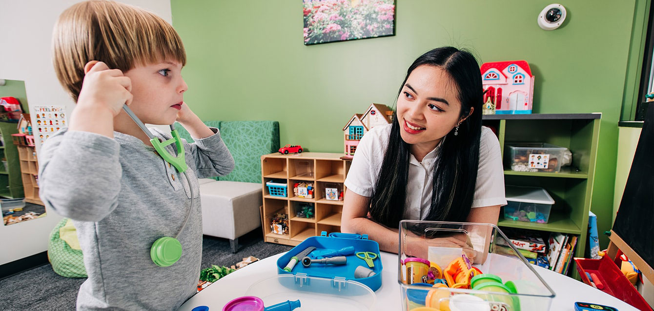 A female student teacher looks on as a young boy tries on a toy stethoscope in a pre-k setting.