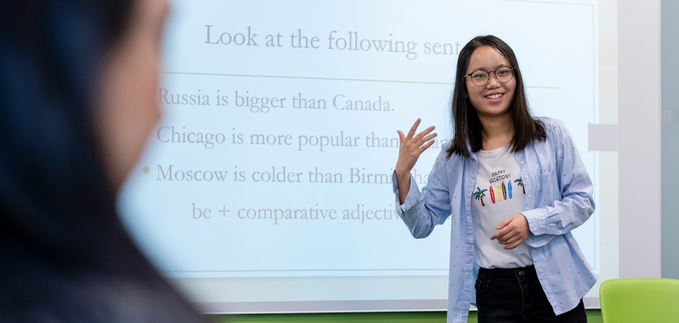 A woman stands in front of a lesson projected on a white baord, gesturing and smiling.