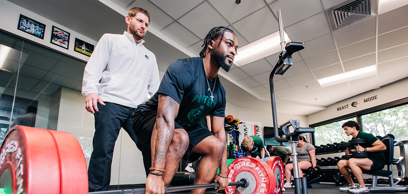 A male trainer watches as a young man squats to lift a heavily weighted barbell.