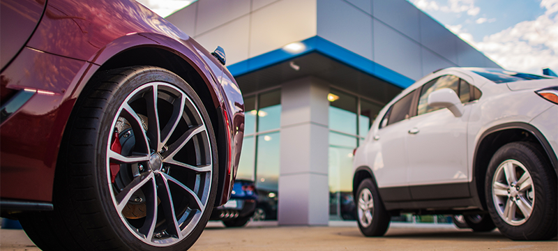 Close-up photo of two new cars taking from below, parked in front of a glass-front car dealership.