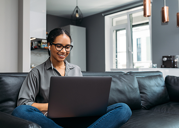 A smiling woman sits cross legged on a leather couch typing on a laptop with earbuds in her ears.