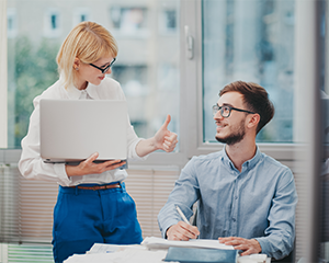 A woman stands over a man's desk giving him a thumbs up