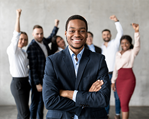 A man in a business suit smiles in the foreground while his direct reports cheer in the background