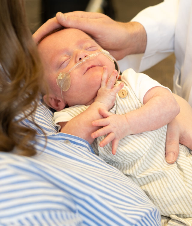 A sleeping infant with a nasal cannula rests in his parent's arms while Dr. Travers gently places a hand on the baby's head.