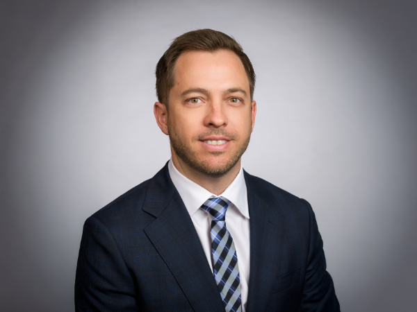 Professional headshot of a man wearing a navy blue suit, white dress shirt, and blue plaid tie, smiling slightly against a light gray background.