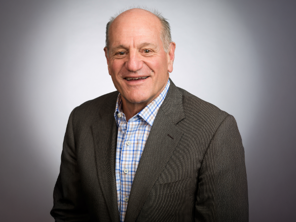 Professional headshot of a smiling man wearing a brown sport coat and blue and orange plaid dress shirt against a gray background.