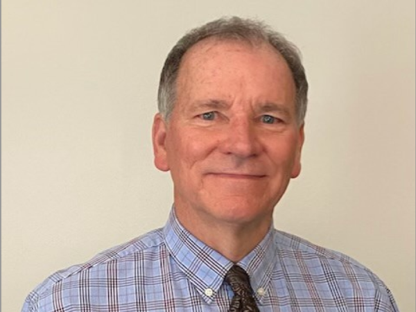 A smiling man in a blue plaid shirt and dark tie stands against a plain background, conveying a professional and approachable demeanor.