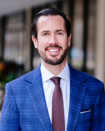 Portrait of a person in a blue suit and tie, with trimmed facial hair, standing outdoors with a blurred background.