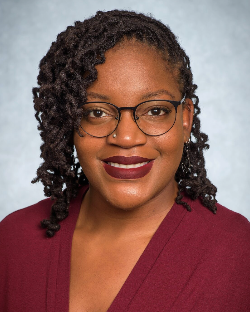 Professional headshot of an individual wearing glasses and a burgundy top against a light gray background.