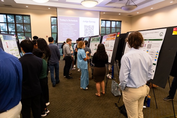 UAB Expo In-Person Poster Presentation in Alumni House, Summer 2024 - Service Learning and Undergraduate Research. Overall of students standing beside their poster presentations and and looking at posters from other students as well, July 2024.