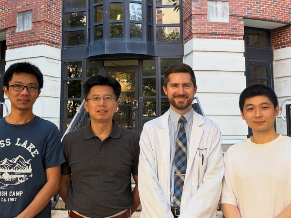 Four individuals stand side by side outdoors in front of a red brick academic building with large windows and stair railings. One person wears a white lab coat and tie, while the others wear casual shirts. The group is centered in front of the building entrance during daylight.