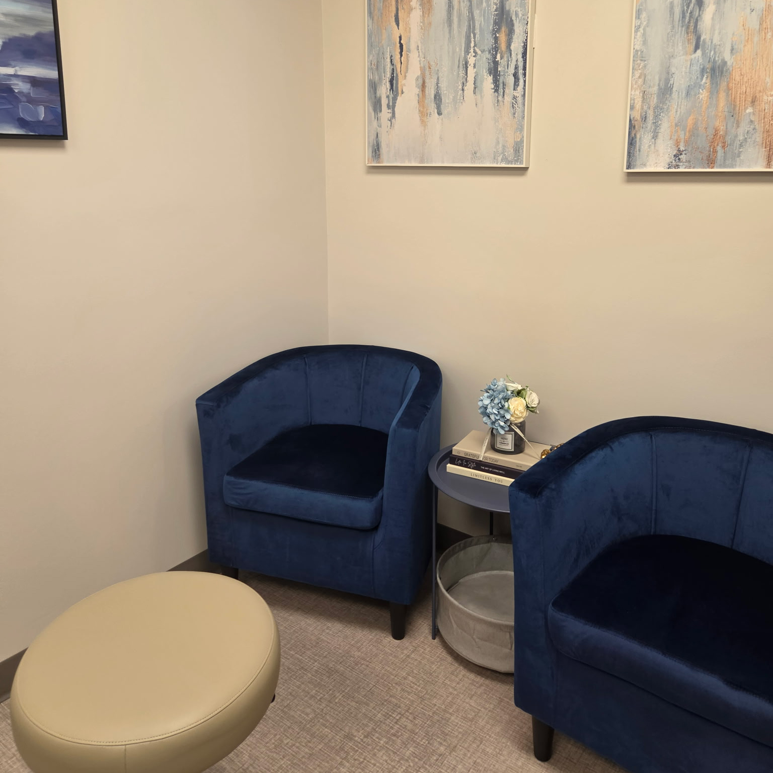 A modern waiting area with two navy blue chairs, a beige ottoman, and abstract wall art.