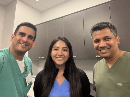 Group photo of three people standing together in a clinical workspace. Two individuals are wearing medical scrubs in green tones, and one person stands between them wearing blue scrubs with a black jacket. All three are smiling toward the camera with cabinets and clinical office furnishings in the background, creating a collegial healthcare team setting.