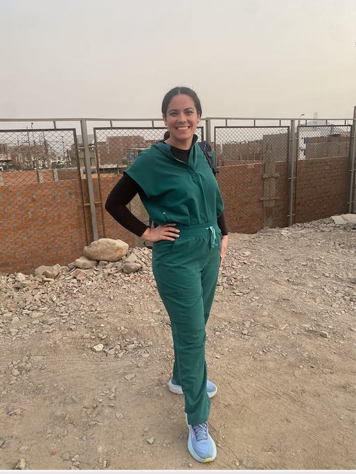 Proenza stands with scrubs up, hand on hip, and smiling, in front of a brick fence with a Peruvian city in the background