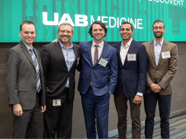 Five attendees pose together in front of a UAB Medicine conference display at a symposium, wearing business attire.