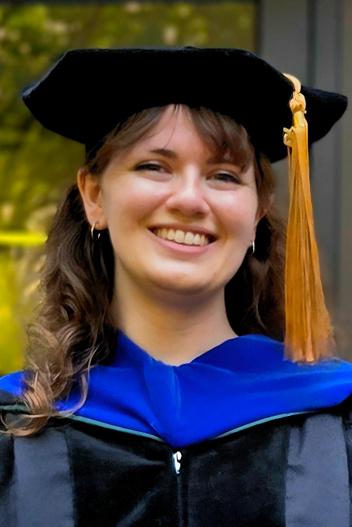 Jennifer Luck, Ph.D., wears a graduation cap and robes while standing outdoors.