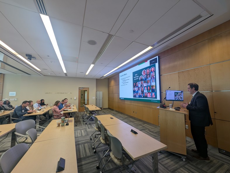 Dr. Mossa-Basha presenting at the front of a room at a podium in front of a screen with a group of faculty, staff, and trainees watching the presentation