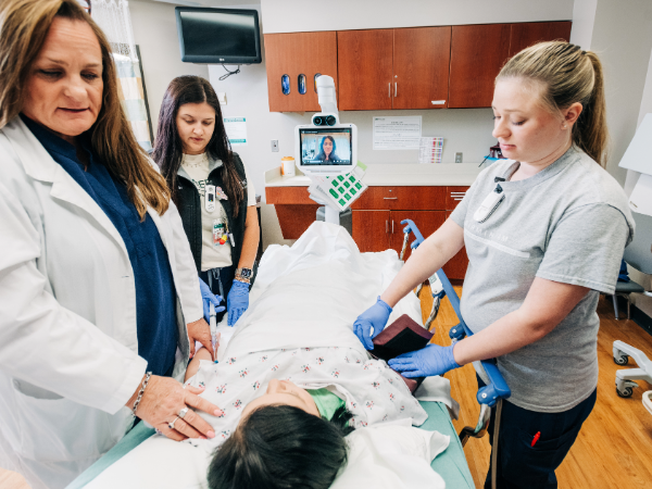 UAB Hospital-Highlands. Three unidentified female healthcare workers are standing beside a hospital bed while speaking with a staged teleneurology (telestroke) female patient in an examination room while Dr. Ekaterina Bakradze, MD (Associate Professor, Neurology) is watching and speaking on a computer screen in the background.