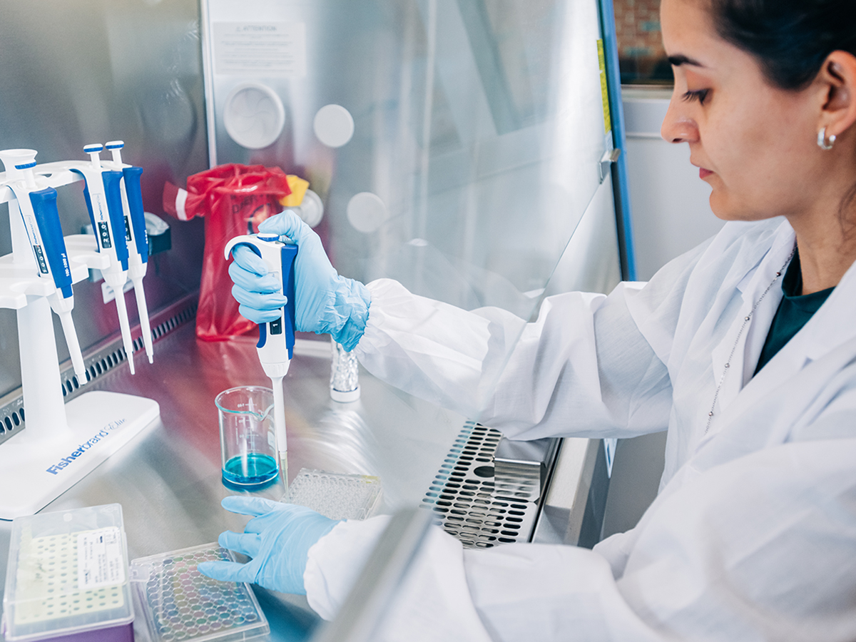 From side, female student wearing lab coat and PPE gloves is using a single-channel pipette while working in a lab exhaust fume hood
