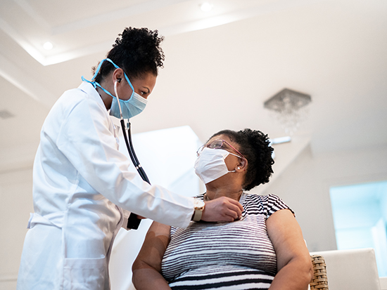 Doctor listening to patient's heartbeat during home visit - wearing face mask