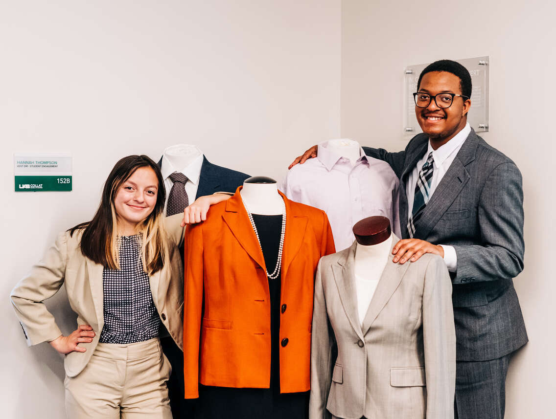 Two smiling students are wearing business suits while standing next to three dressmaker's mannequins clothed in male and female business suit