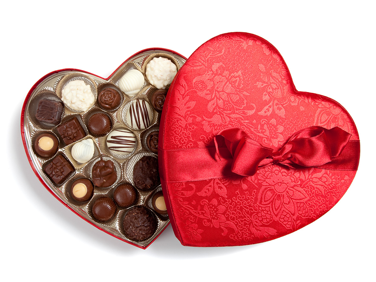 A red heart shaped boxed of chocolates that is open on a white back ground displaying the exterior and interior of the box 