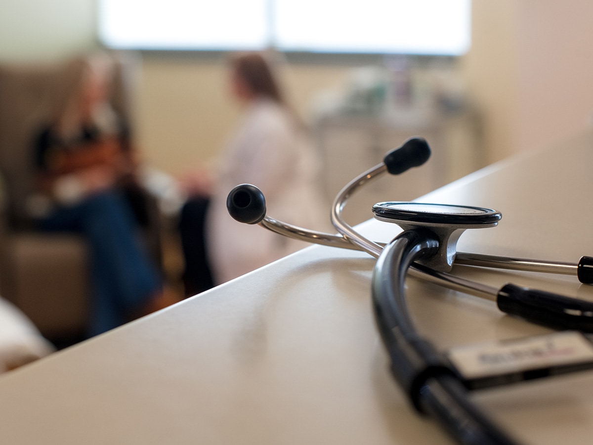 Stethoscope resting on counter with two blurred people talking in a clinical setting in the background. 