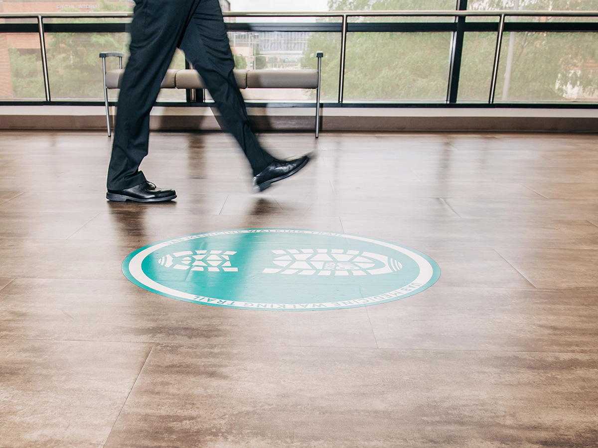 Close-up, action shot of legs and feet of person walking on floor signage decal that reads "UAB Medicine Walking Trail" in UAB Hospital, May 2025.