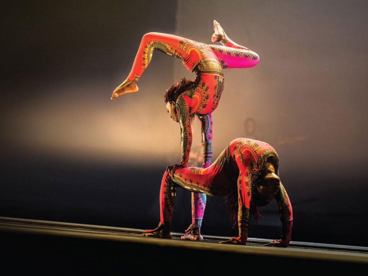 Performers in Cirque Kalabanté perform acrobatic weight-sharing stunts on stage dressed in brightly colored costumes.