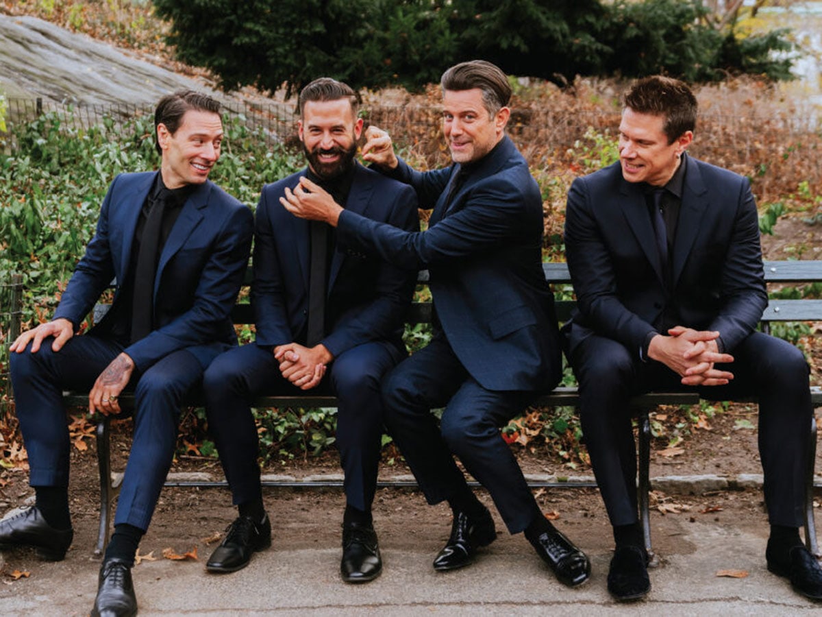 Members of the group Il Divo pose together in suits while sitting on a bench outdoors.