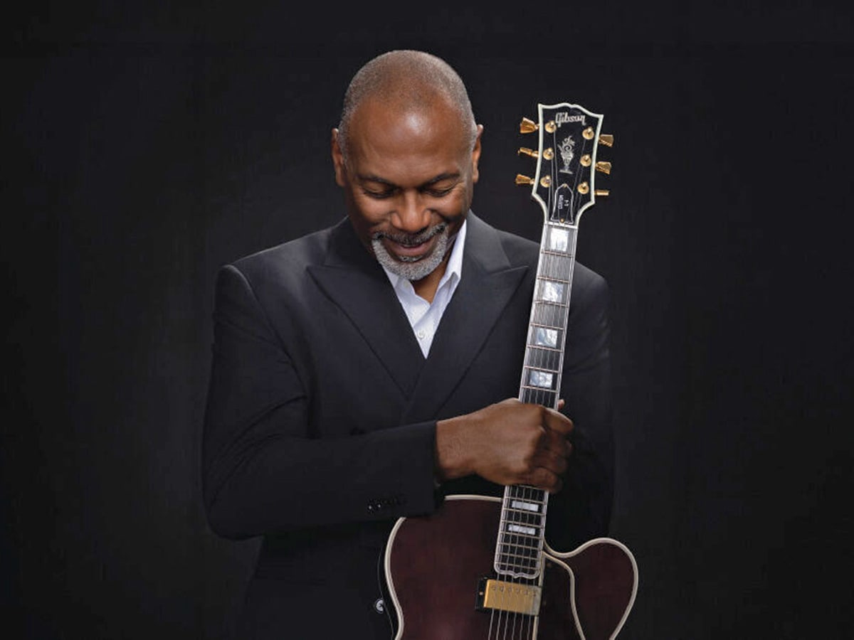 Jazz musician Eric Essix wears a dark suit and holds his guitar over his heart, posing in front of a dark backdrop.)