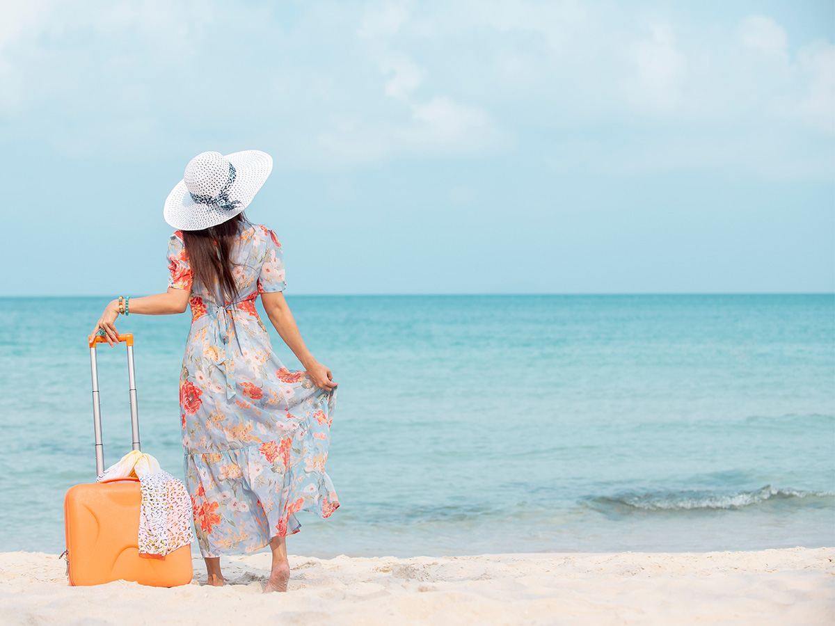 Woman standing on the sand looking out into the ocean wearing a blue floral dress and sun hat while holding an orange suitcase.