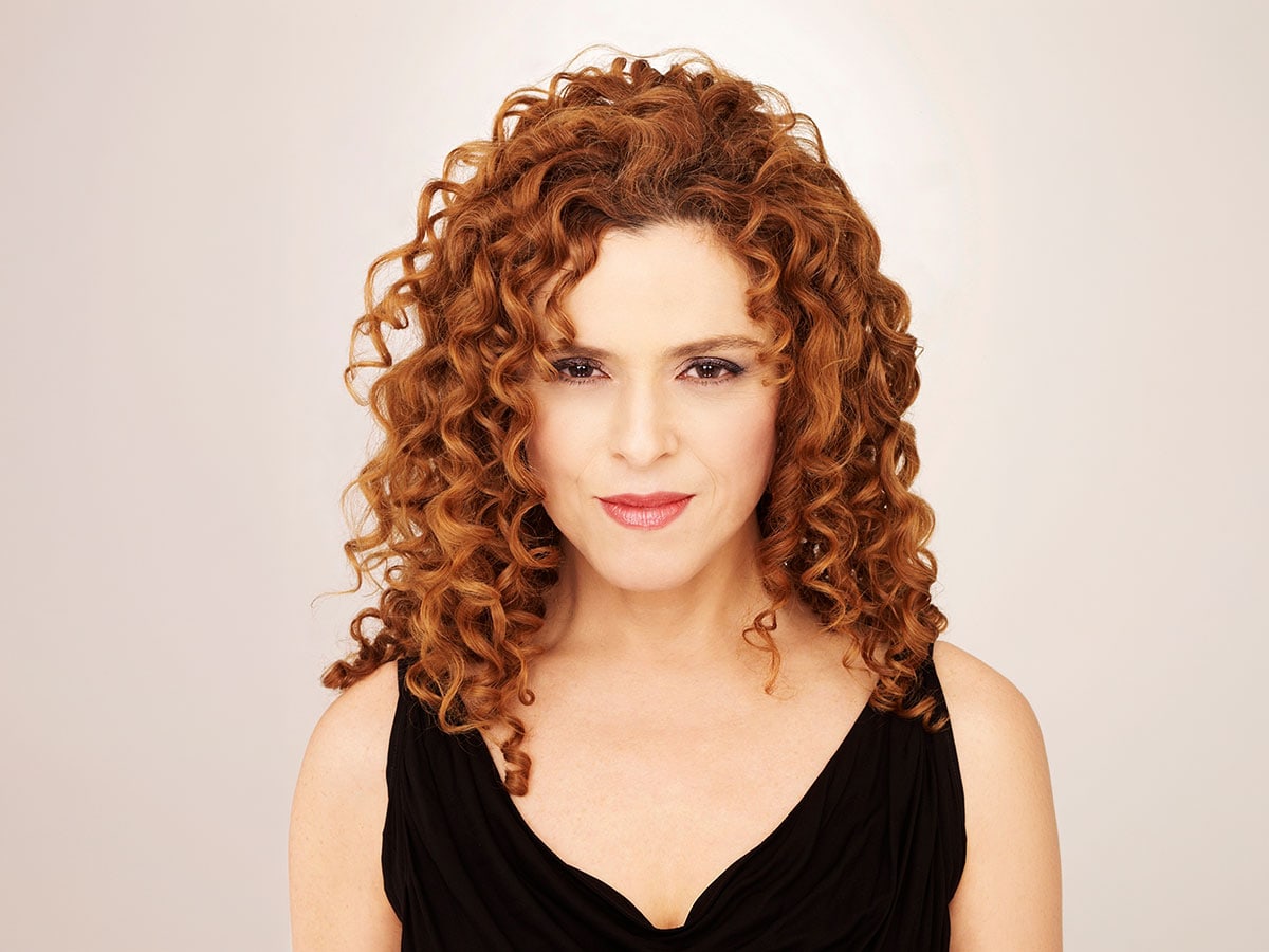 A woman with red curly hair standing against a light background smiles at the camera
