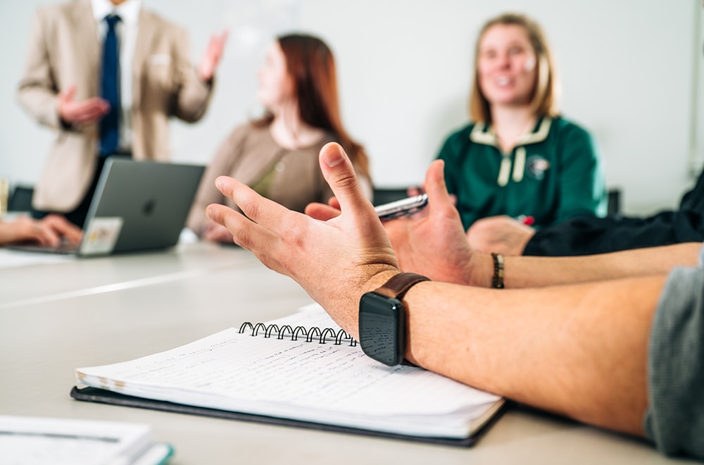 Closeup on an employee's hands, raised in a gesture while making a point, with three other employees visible around a meeting table in the background, their faces blurred
