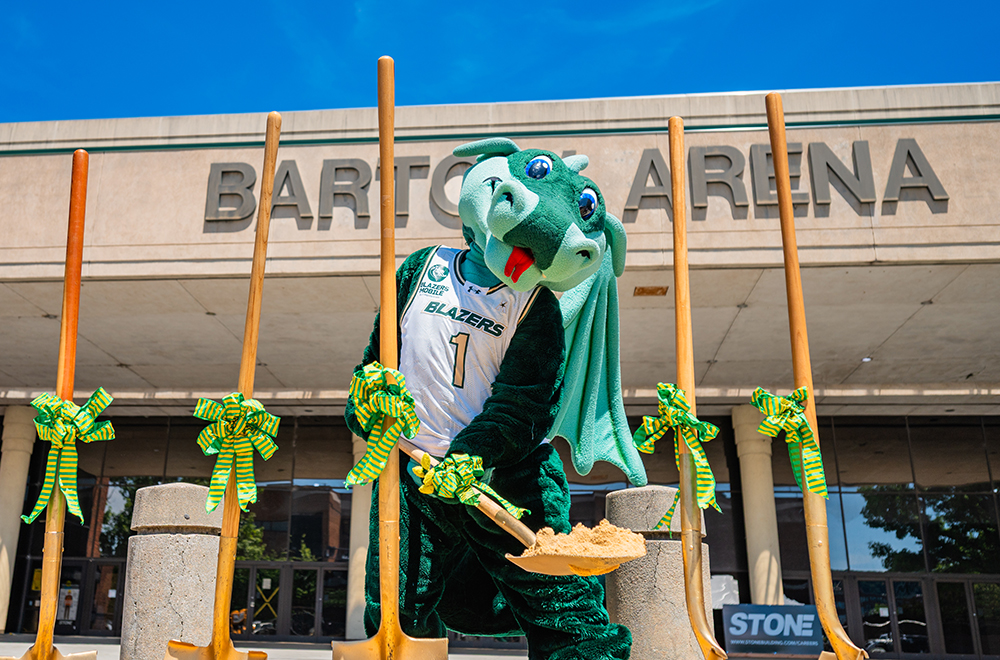 U A B mascot Blaze lifts a spade full of dirt while standing in front of Bartow Arena while other gold-painted shovels, with green bows, are planted in the dirt surrounding Blaze
